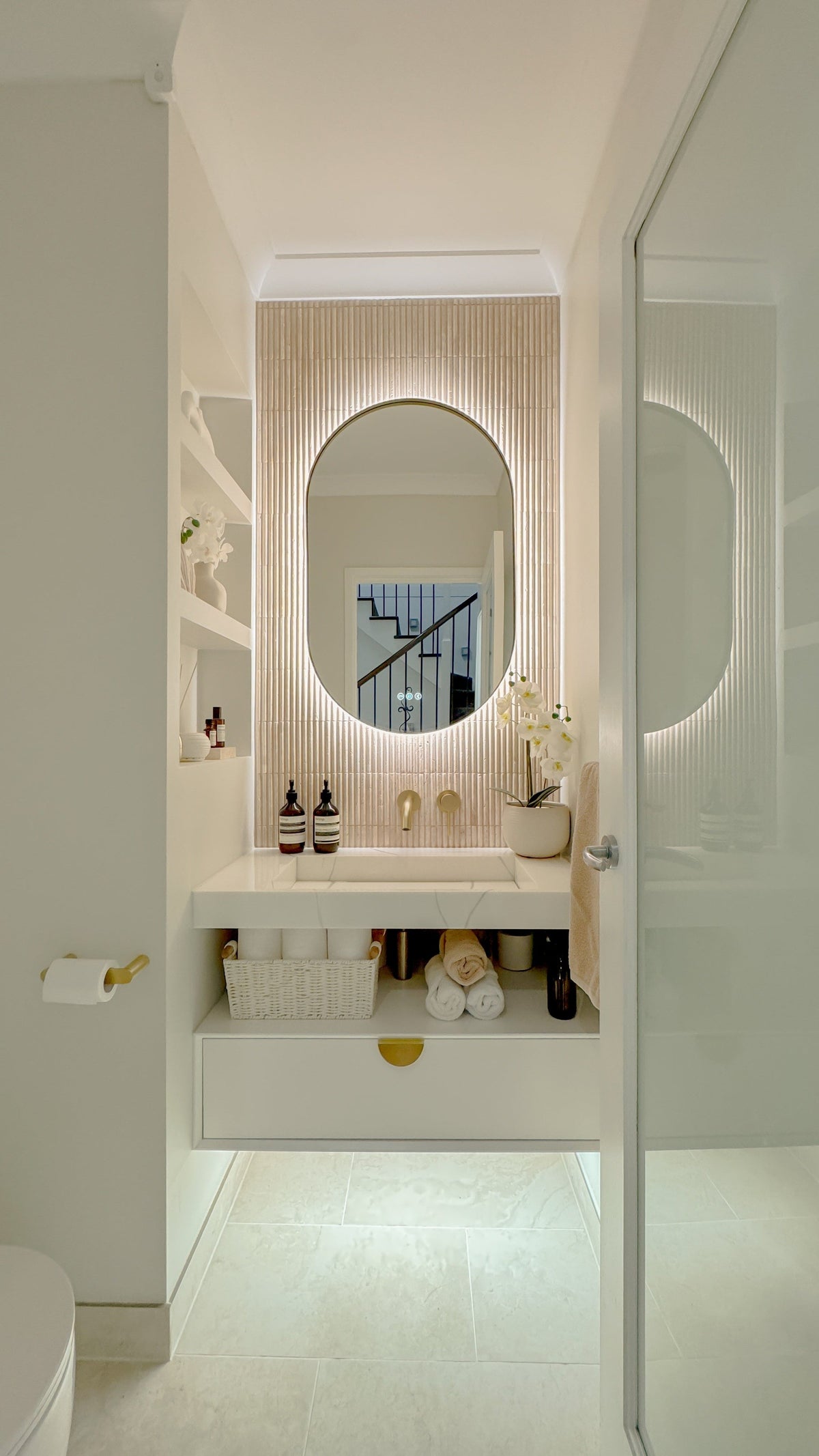 The Valencia 600 x 1000 mm oval framed backlit LED mirror in Brushed Brass, seen through the doorway of a calm white powder room