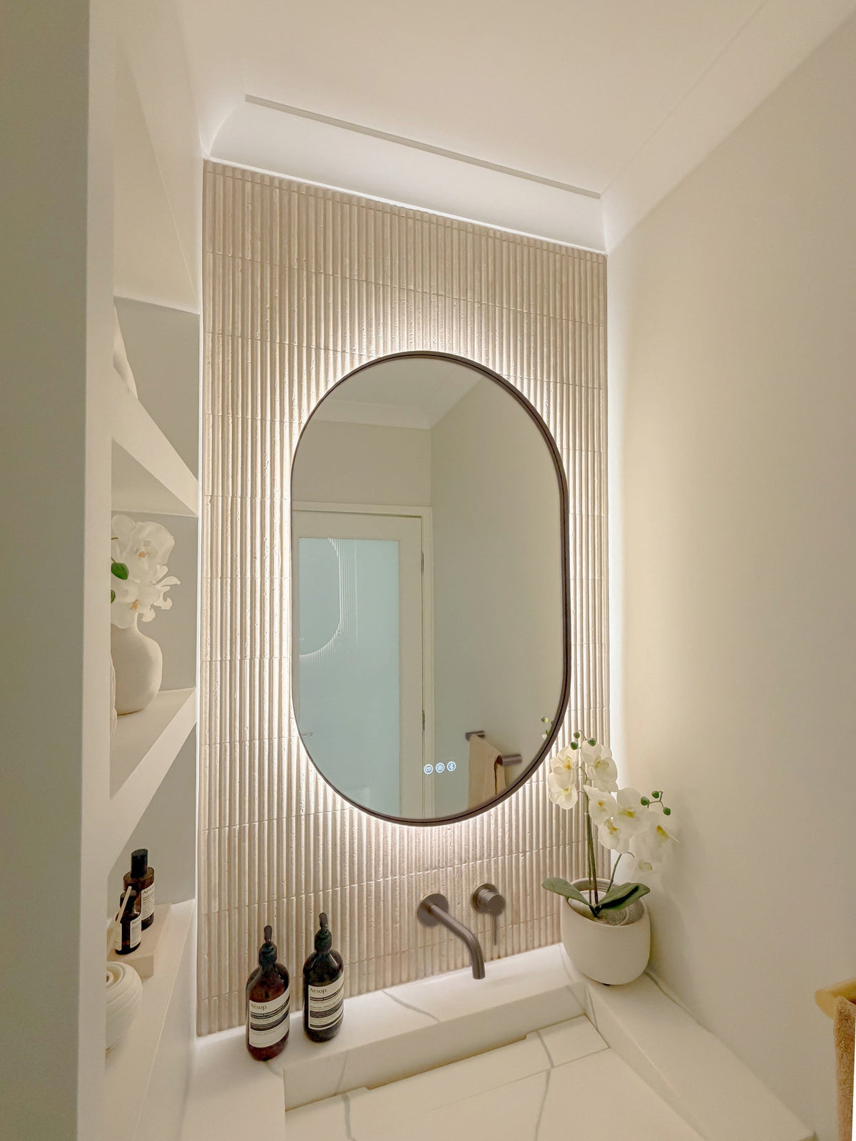 The Valencia 600 x 1000 mm oval framed backlit LED mirror in Antique Brass, seen from the side on a backlit fluted travertine wall, showing the slim frame edge