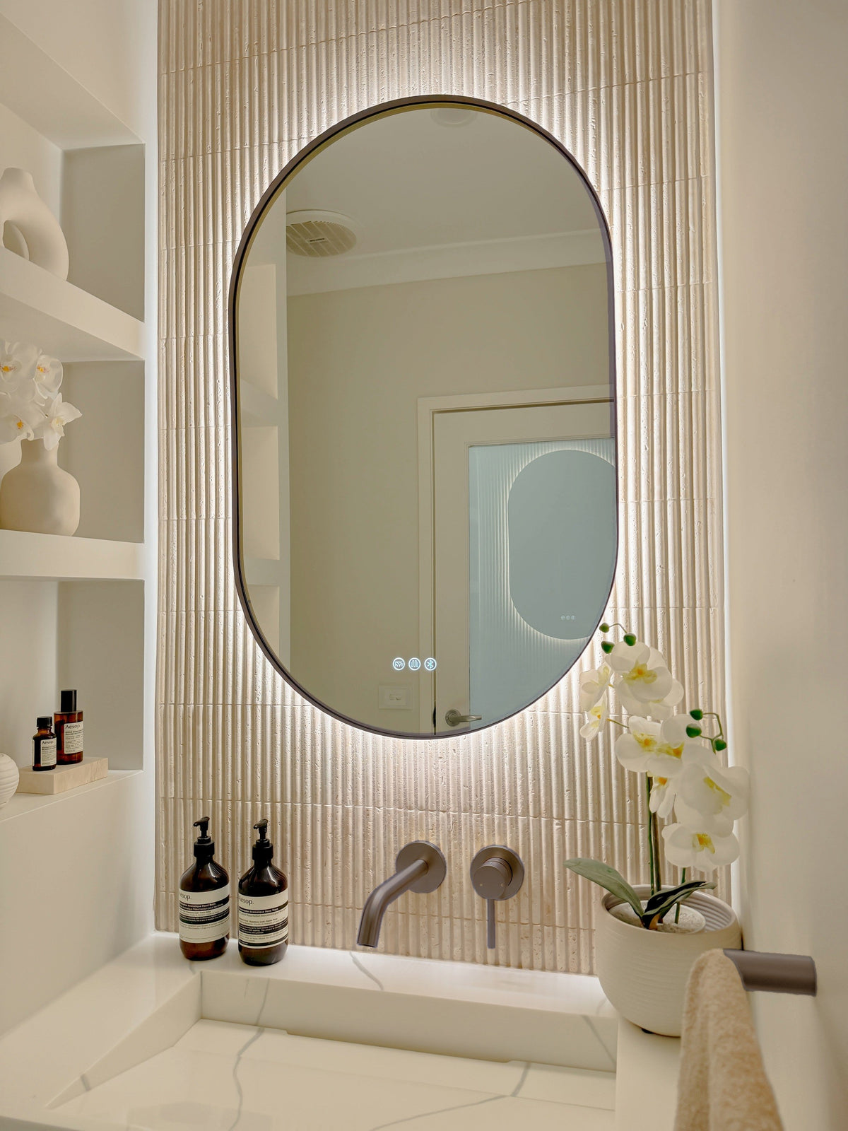 The Valencia 600 x 1000 mm oval framed backlit LED mirror in Antique Brass, angled close view with the warm backlight glowing on fluted travertine