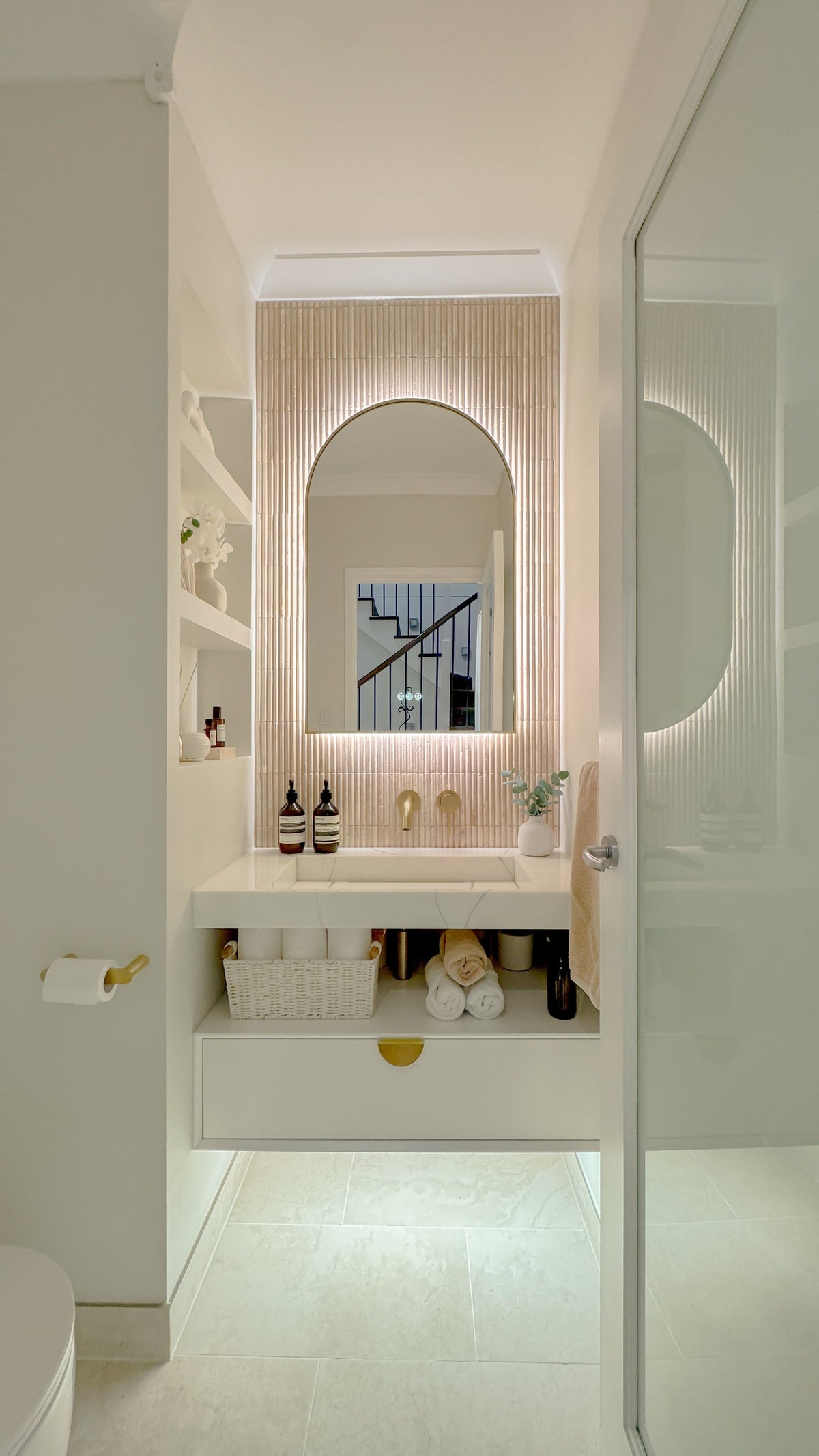 The Marbella 600 x 900 mm arch framed backlit LED mirror in Brushed Brass, seen through the doorway of a calm white powder room