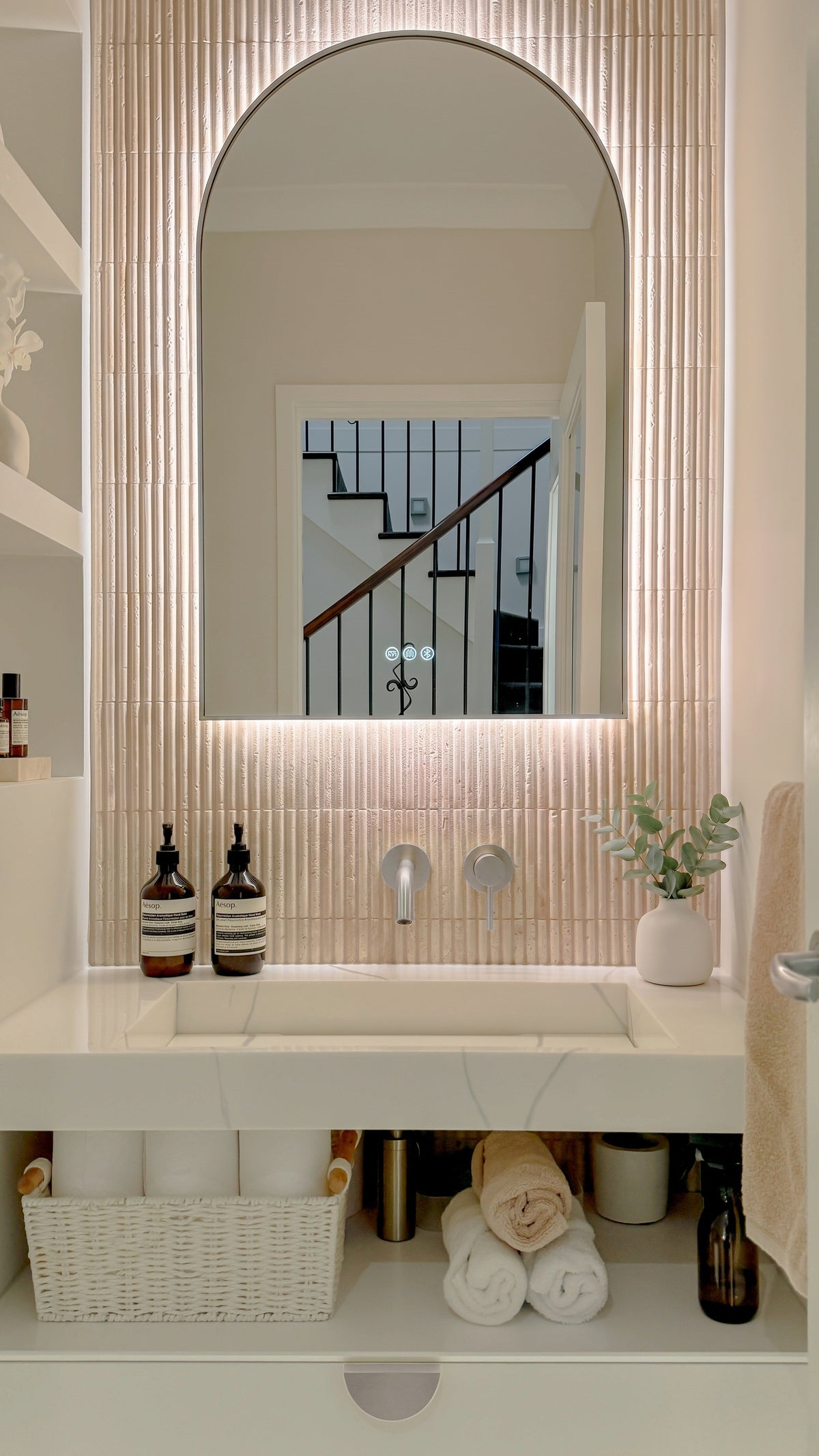 The Marbella 600 x 1000 mm arch framed backlit LED mirror in Brushed Nickel, above a white stone basin with a matching Milano wall spout and mixer, fluted travertine wall