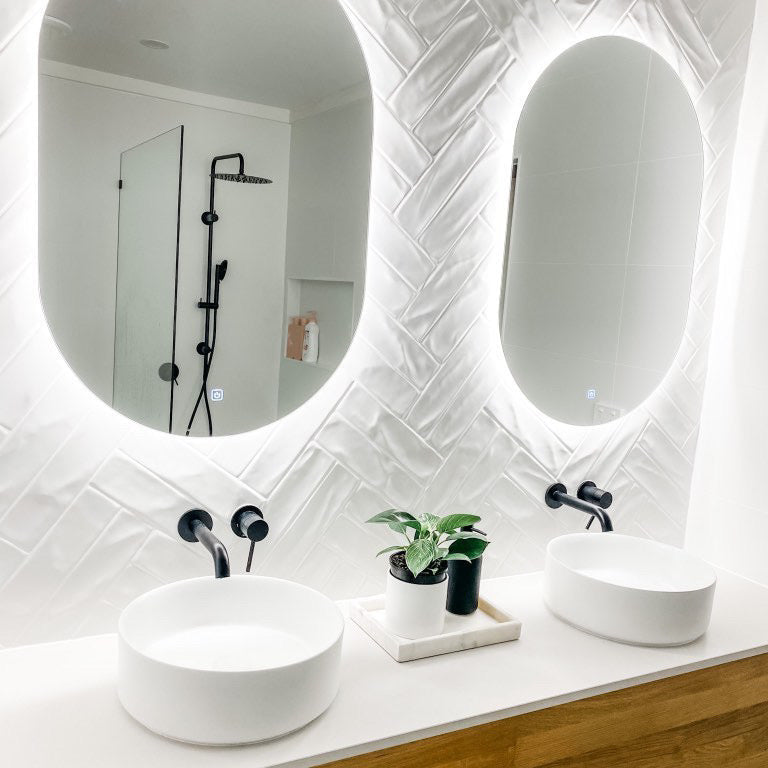 Bright vanity area with dual Oval LED mirrors and floating cabinet on white herringbone tiled wall