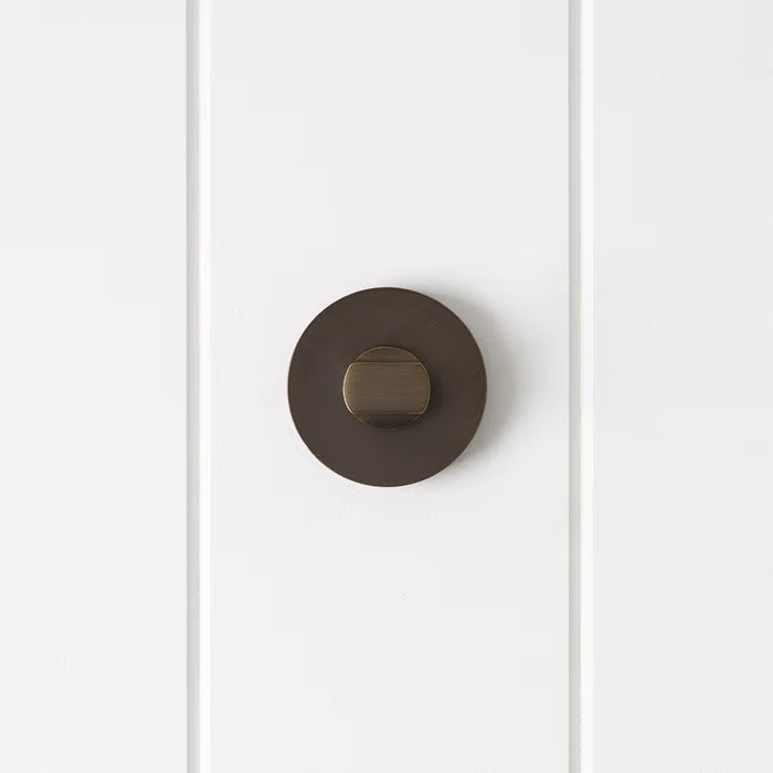 Prague Door Lock in Antique Brass, front view of the thumb-turn on its round rose, white vertical-panelled wall