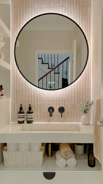 The Capri round 800 mm framed backlit LED mirror in Matte Black, above a white stone basin with a matching Milano wall spout and mixer, fluted travertine wall
