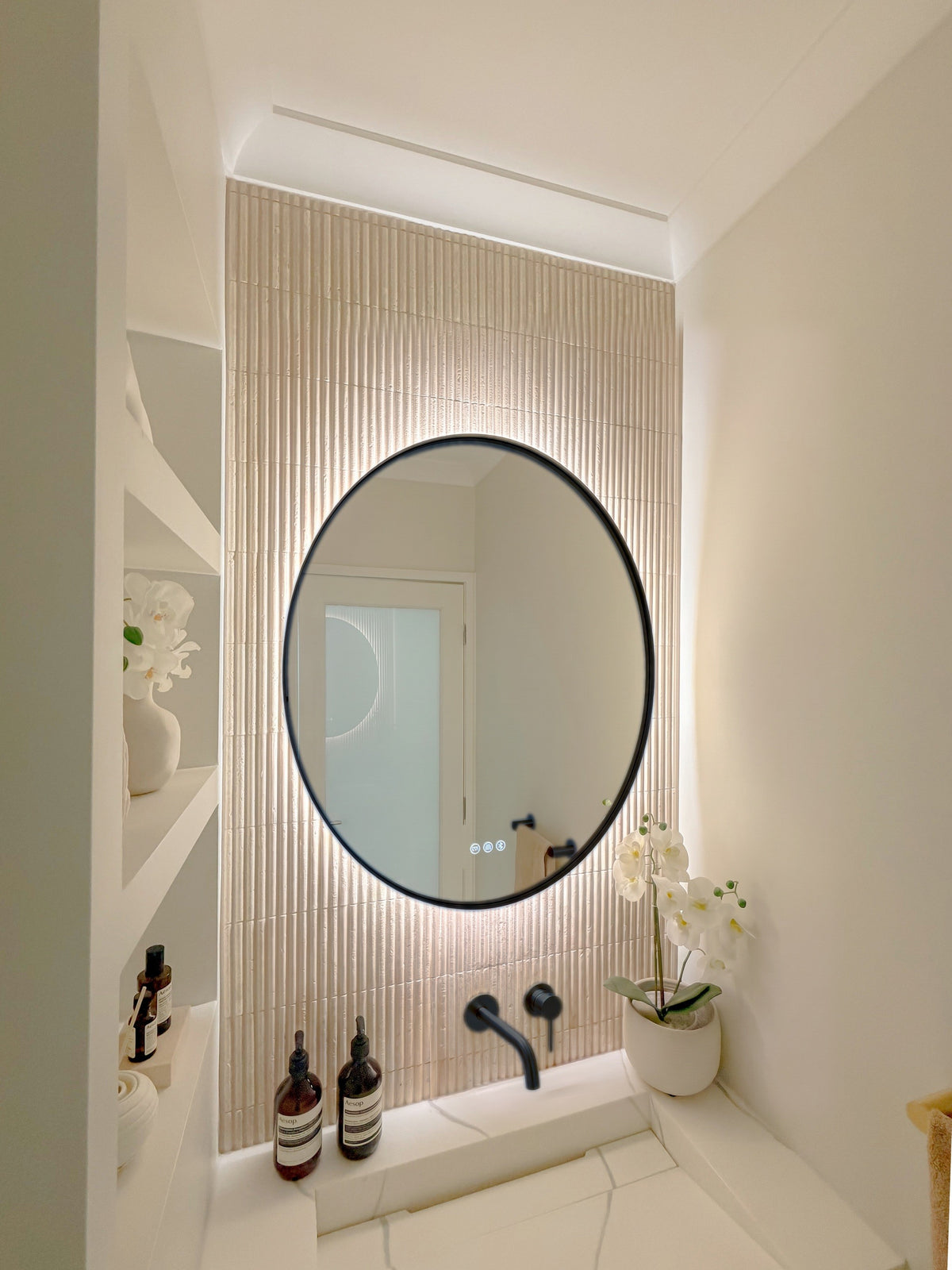 The Capri round 800 mm framed backlit LED mirror in Matte Black, seen from the side on a backlit fluted travertine wall, showing the slim frame edge