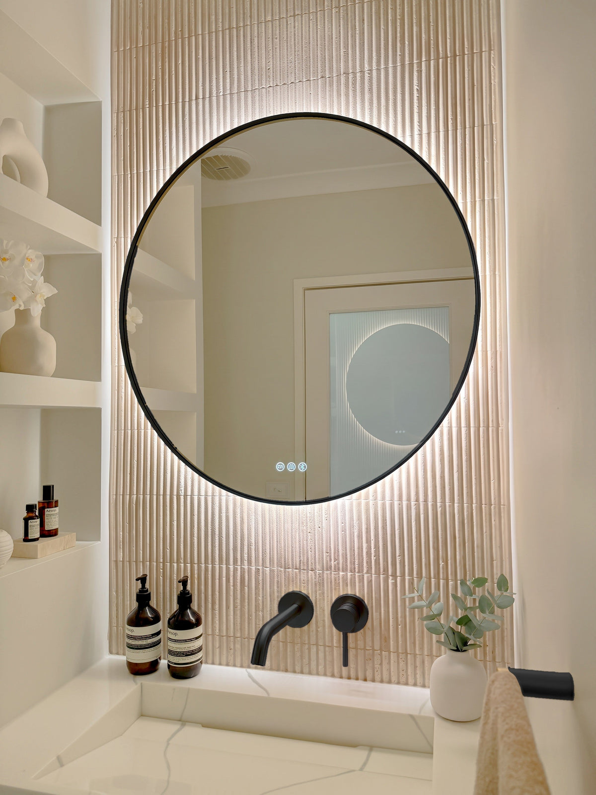 The Capri round 800 mm framed backlit LED mirror in Matte Black, angled close view with the warm backlight glowing on fluted travertine