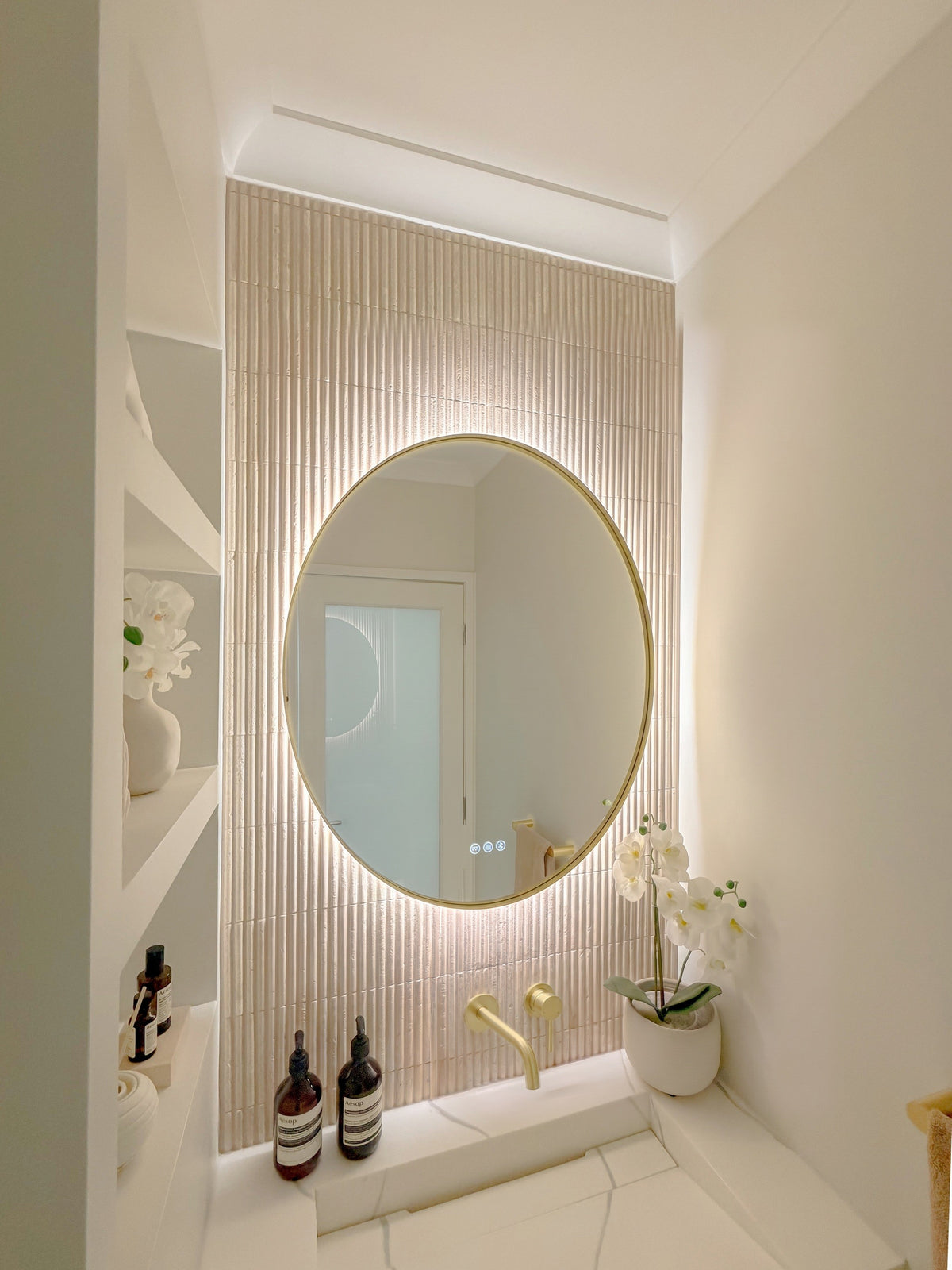 The Capri round 800 mm framed backlit LED mirror in Brushed Brass, seen from the side on a backlit fluted travertine wall, showing the slim frame edge