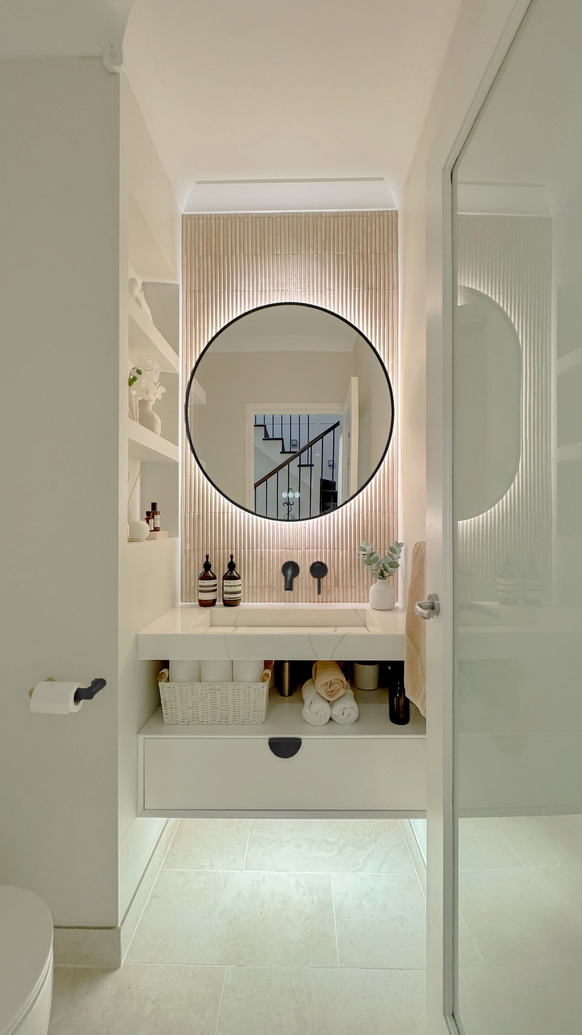 The Capri round 800 mm framed backlit LED mirror in Matte Black, seen through the doorway of a calm white powder room