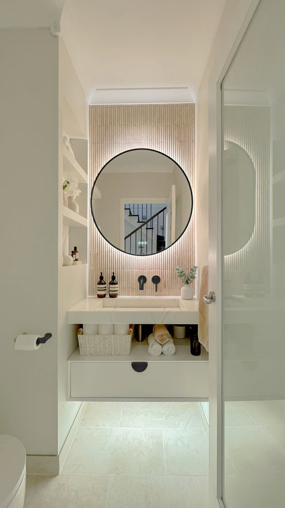 The Capri round 800 mm framed backlit LED mirror in Matte Black, seen through the doorway of a calm white powder room