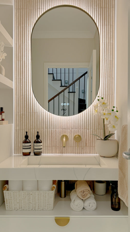 The Valencia 600 x 900 mm oval framed backlit LED mirror in Brushed Brass, above a white stone basin with a matching Milano wall spout and mixer, fluted travertine wall