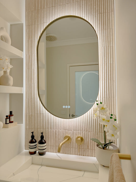 The Valencia 600 x 900 mm oval framed backlit LED mirror in Brushed Brass, angled close view with the warm backlight glowing on fluted travertine