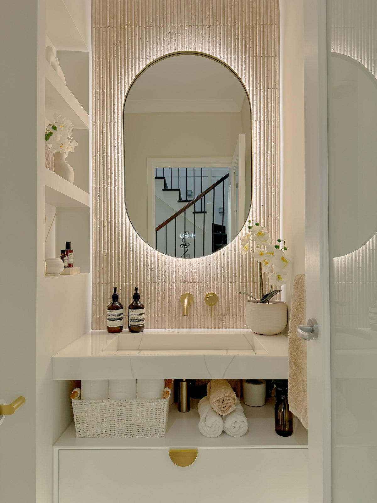The Valencia 600 x 1000 mm oval framed backlit LED mirror in Brushed Brass, in a powder room above a floating white vanity with matching hardware