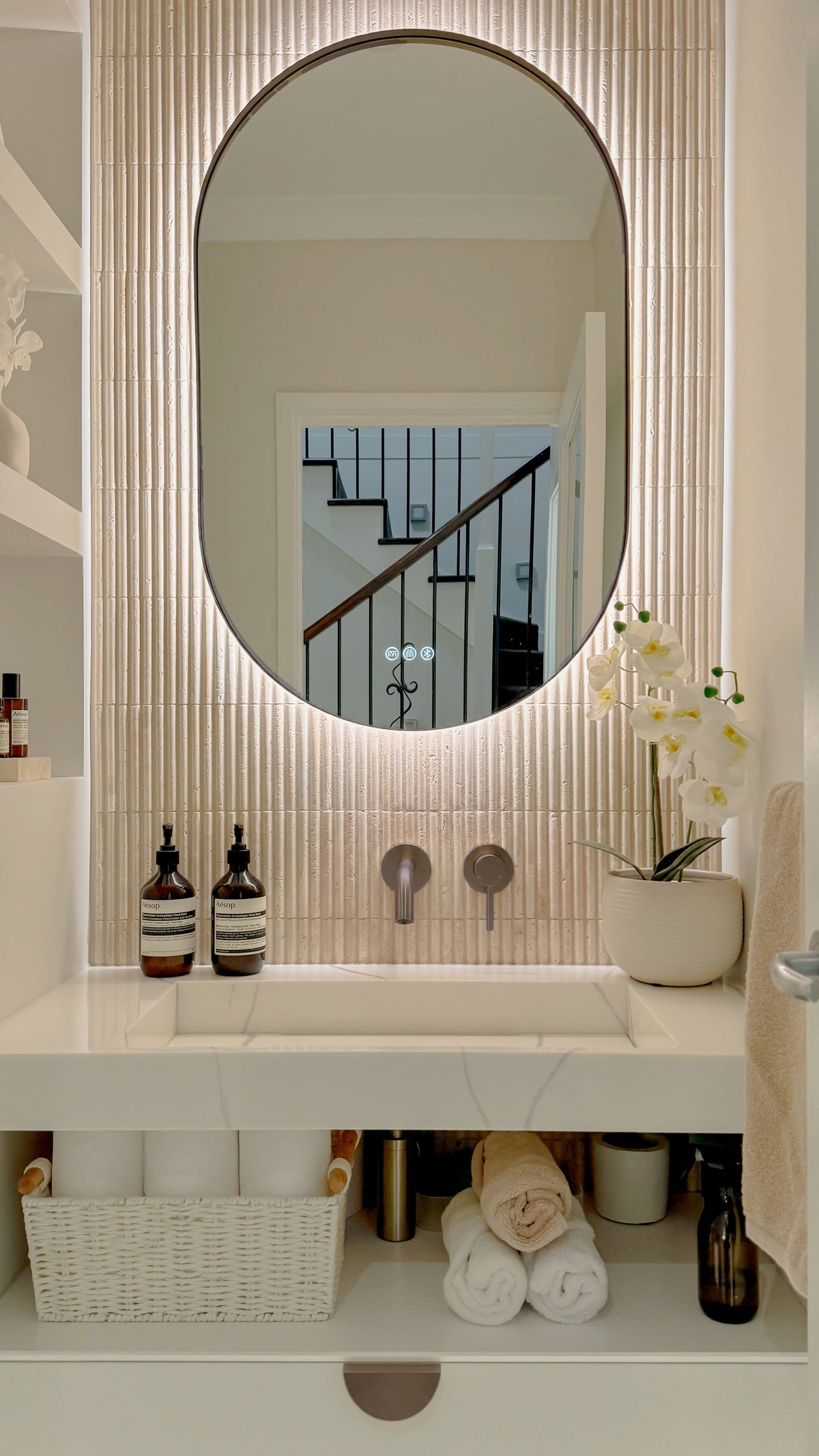 The Valencia 600 x 1000 mm oval framed backlit LED mirror in Antique Brass, above a white stone basin with a matching Milano wall spout and mixer, fluted travertine wall