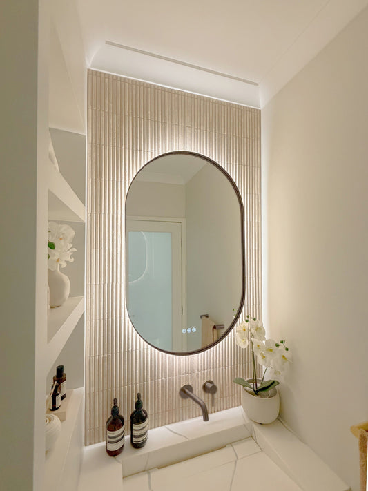 The Valencia 600 x 900 mm oval framed backlit LED mirror in Antique Brass, seen from the side on a backlit fluted travertine wall, showing the slim frame edge