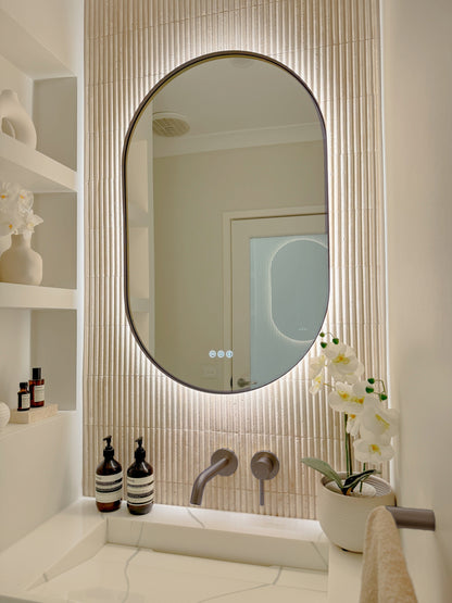 The Valencia 600 x 1000 mm oval framed backlit LED mirror in Antique Brass, angled close view with the warm backlight glowing on fluted travertine