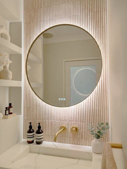 The Capri round 800 mm framed backlit LED mirror in Brushed Brass, angled close view with the warm backlight glowing on fluted travertine