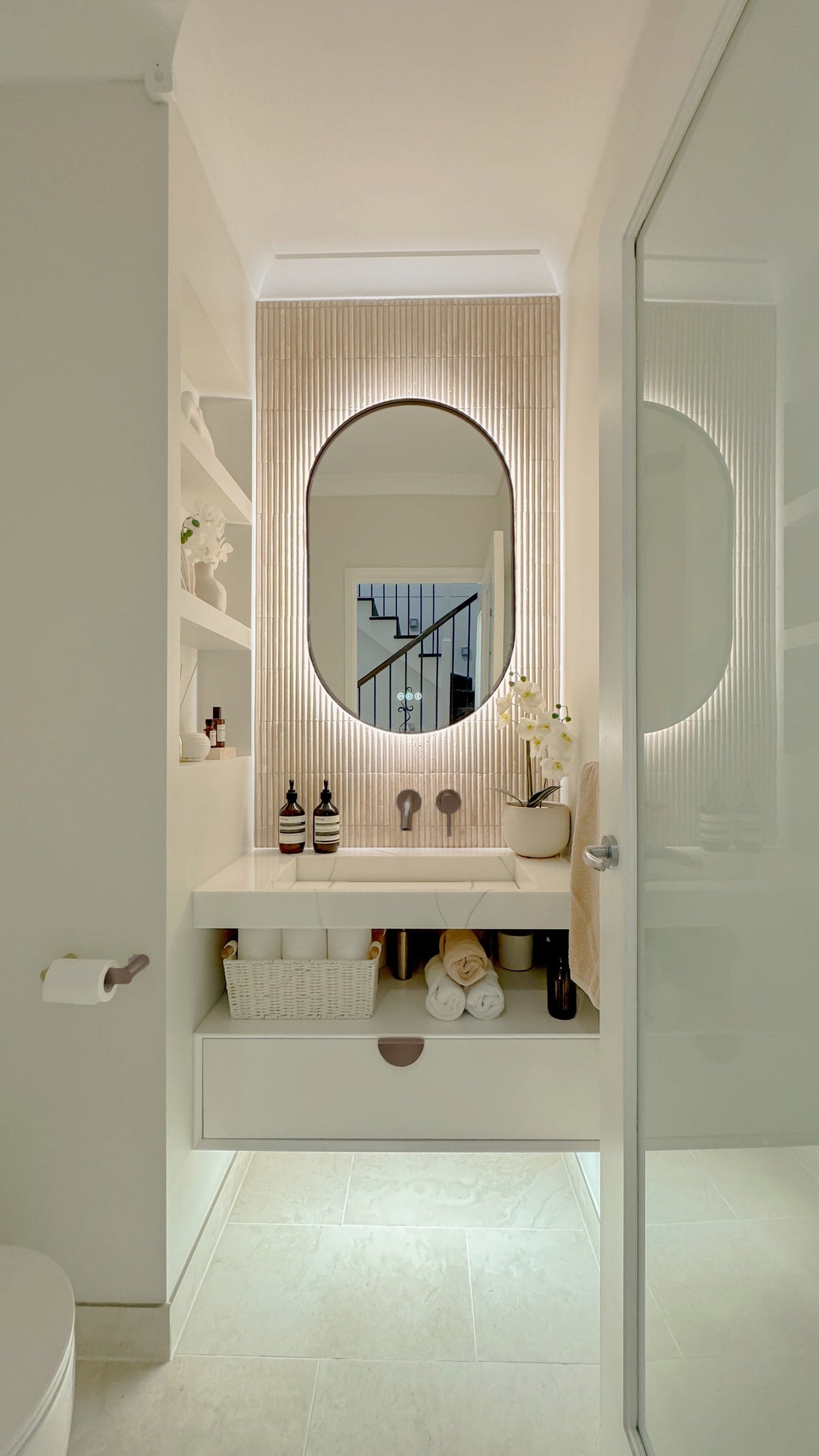 The Valencia 600 x 1000 mm oval framed backlit LED mirror in Antique Brass, seen through the doorway of a calm white powder room
