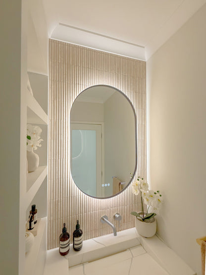 The Valencia 600 x 1000 mm oval framed backlit LED mirror in Brushed Nickel, seen from the side on a backlit fluted travertine wall, showing the slim frame edge