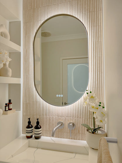 The Valencia 600 x 1000 mm oval framed backlit LED mirror in Brushed Nickel, angled close view with the warm backlight glowing on fluted travertine