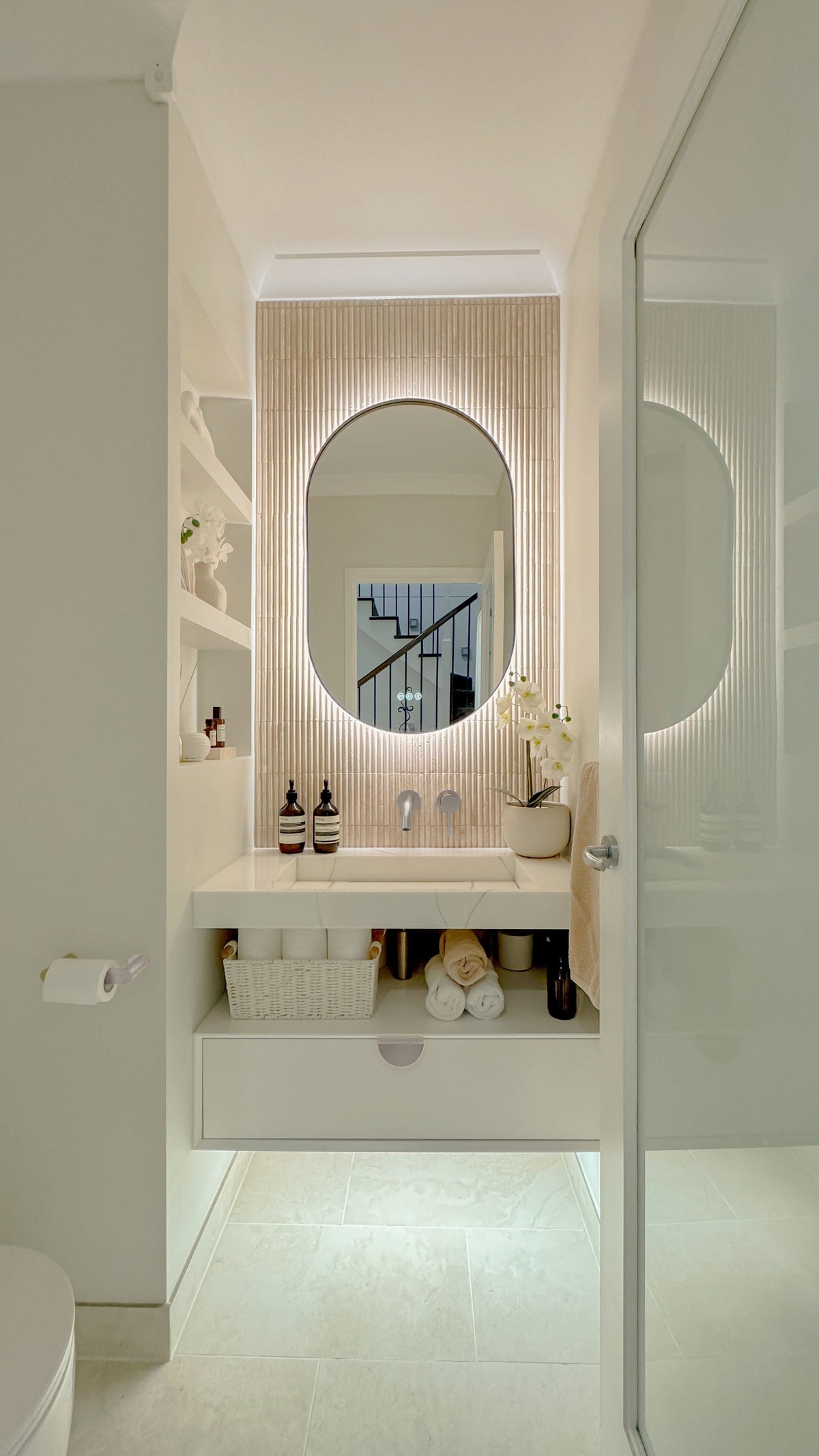 The Valencia 600 x 1000 mm oval framed backlit LED mirror in Brushed Nickel, seen through the doorway of a calm white powder room