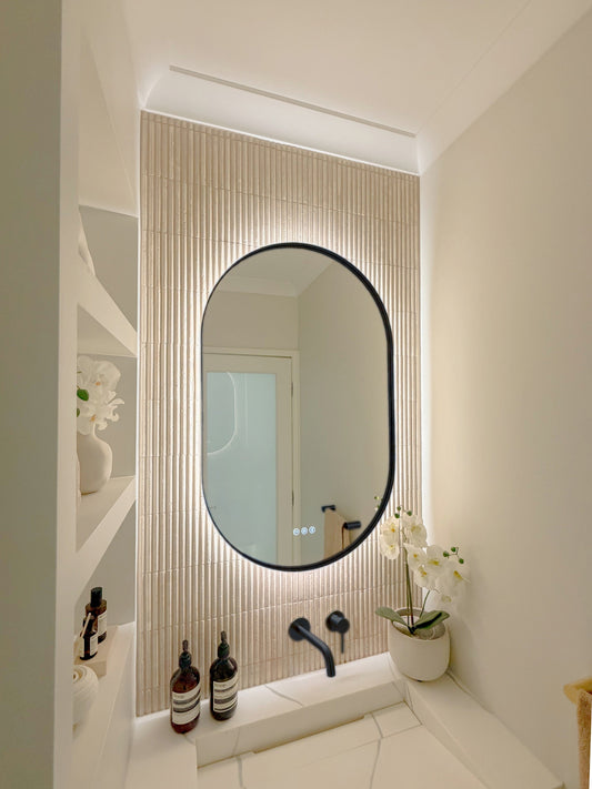 The Valencia 600 x 900 mm oval framed backlit LED mirror in Matte Black, seen from the side on a backlit fluted travertine wall, showing the slim frame edge