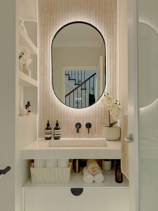 The Valencia 600 x 1000 mm oval framed backlit LED mirror in Matte Black, in a powder room above a floating white vanity with matching hardware