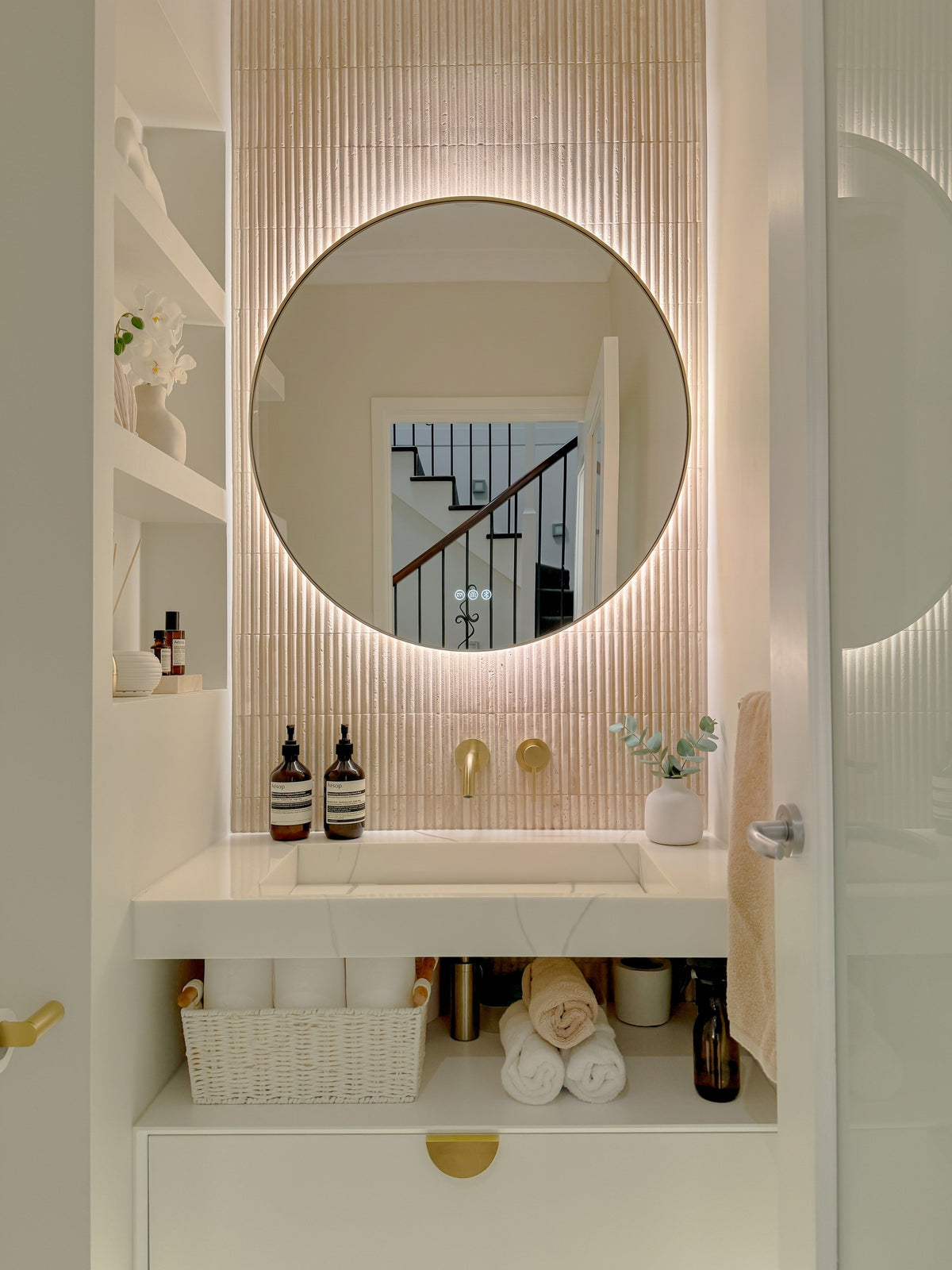 The Capri round 800 mm framed backlit LED mirror in Brushed Brass, in a powder room above a floating white vanity with matching hardware
