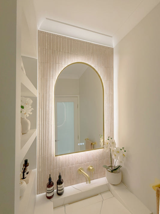The Marbella 600 x 900 mm arch framed backlit LED mirror in Brushed Brass, seen from the side on a backlit fluted travertine wall, showing the slim frame edge