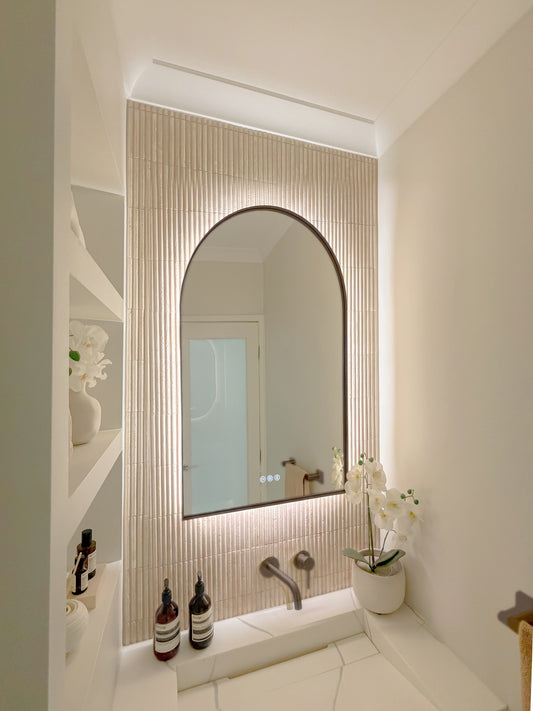The Marbella 600 x 1000 mm arch framed backlit LED mirror in Antique Brass, seen from the side on a backlit fluted travertine wall, showing the slim frame edge