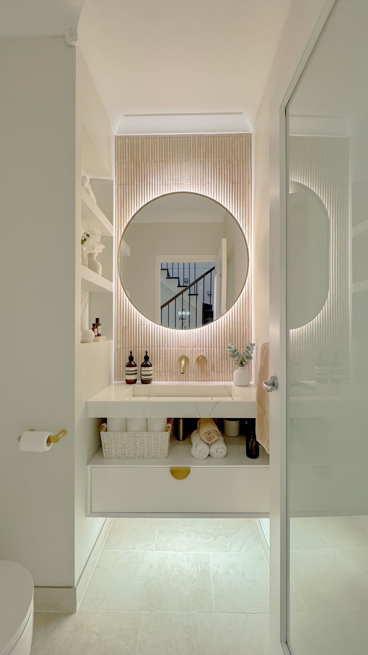 The Capri round 800 mm framed backlit LED mirror in Brushed Brass, seen through the doorway of a calm white powder room