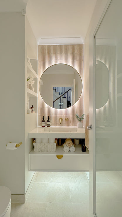 The Capri round 800 mm framed backlit LED mirror in Brushed Brass, seen through the doorway of a calm white powder room