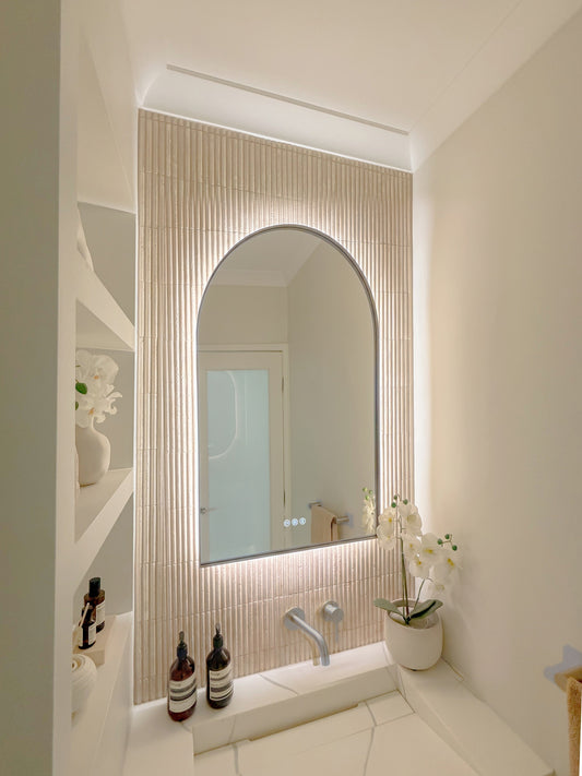 The Marbella 600 x 900 mm arch framed backlit LED mirror in Brushed Nickel, seen from the side on a backlit fluted travertine wall, showing the slim frame edge