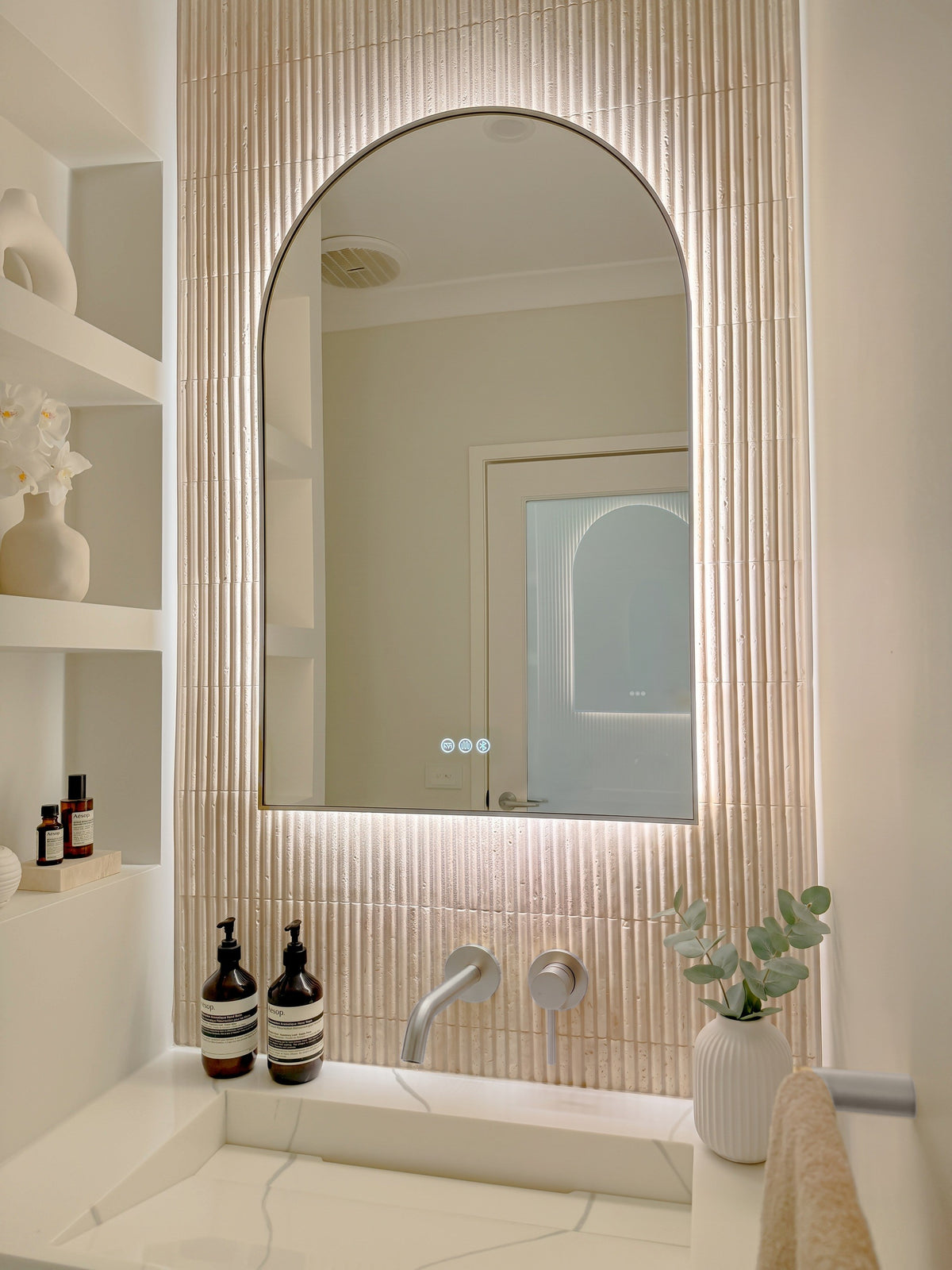 The Marbella 600 x 1000 mm arch framed backlit LED mirror in Brushed Nickel, angled close view with the warm backlight glowing on fluted travertine