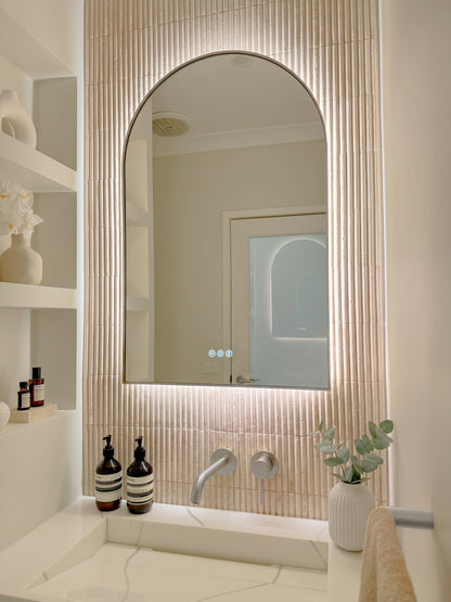 The Marbella 600 x 1000 mm arch framed backlit LED mirror in Brushed Nickel, angled close view with the warm backlight glowing on fluted travertine