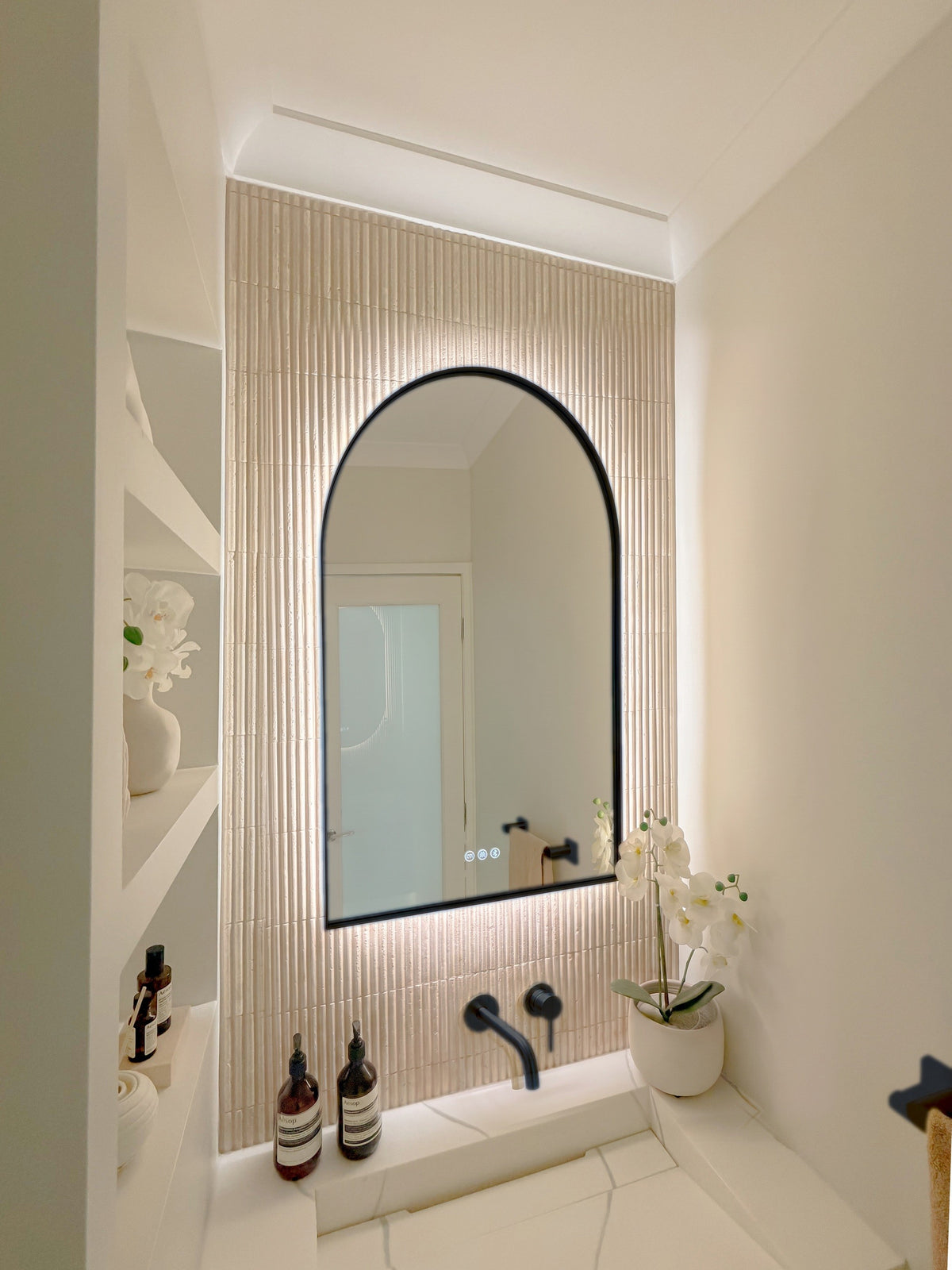 The Marbella 600 x 900 mm arch framed backlit LED mirror in Matte Black, seen from the side on a backlit fluted travertine wall, showing the slim frame edge