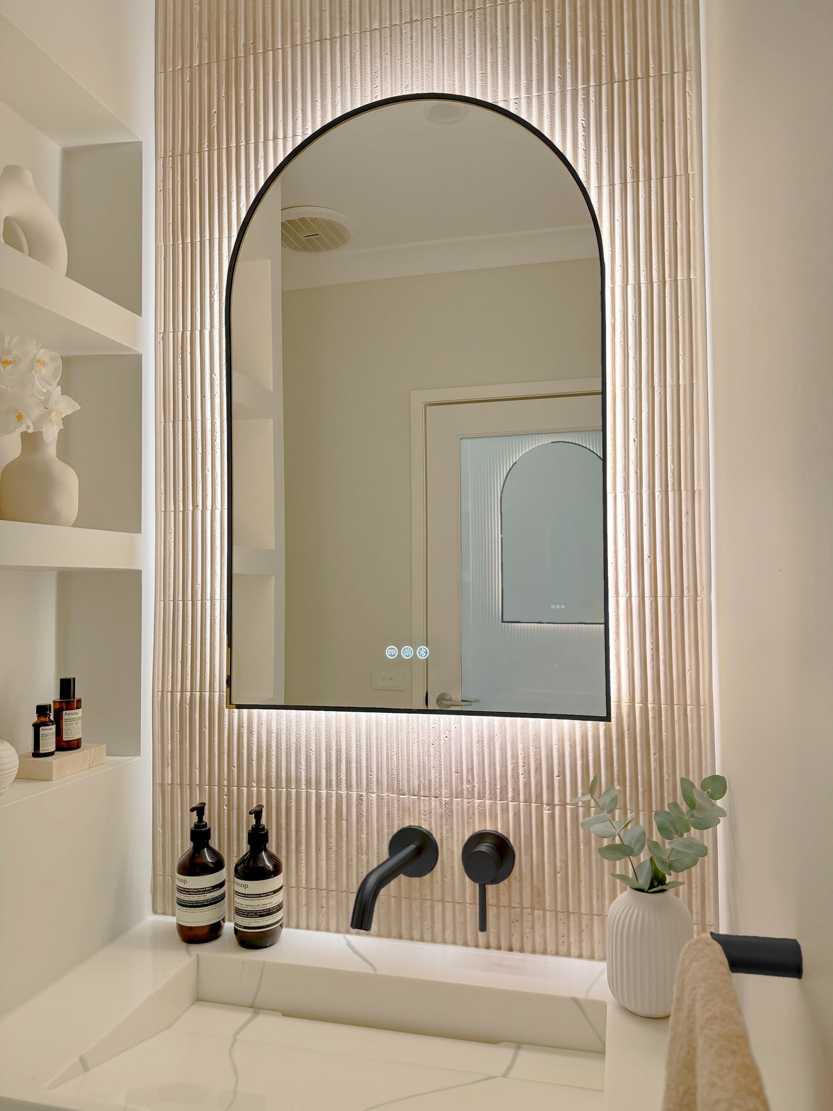 The Marbella 600 x 900 mm arch framed backlit LED mirror in Matte Black, angled close view with the warm backlight glowing on fluted travertine