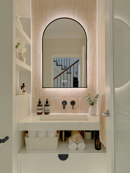 The Marbella 600 x 900 mm arch framed backlit LED mirror in Matte Black, in a powder room above a floating white vanity with matching hardware