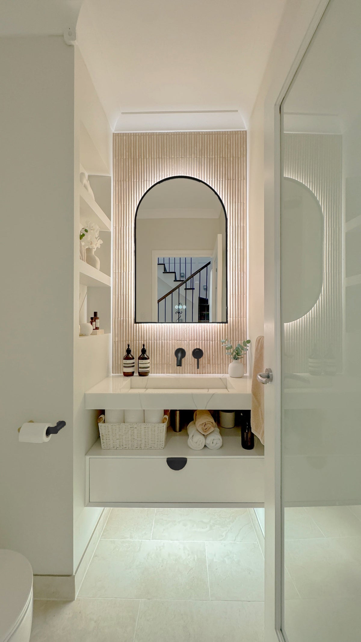 The Marbella 600 x 900 mm arch framed backlit LED mirror in Matte Black, seen through the doorway of a calm white powder room