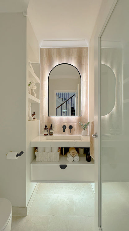 The Marbella 600 x 900 mm arch framed backlit LED mirror in Matte Black, seen through the doorway of a calm white powder room