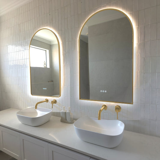 Two Calais arch-shaped LED mirrors with gold wrap-around frames above twin vessel basins with brushed brass tapware