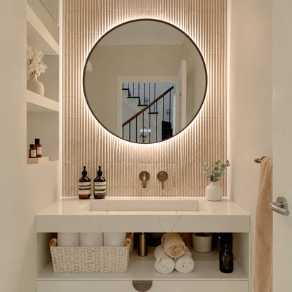 The Capri round 800 mm framed backlit LED mirror in Antique Brass, lit and seen straight-on above a white stone basin with a matching wall spout and mixer