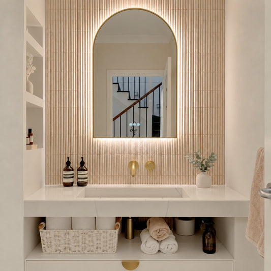 The Marbella 600 x 1000 mm arch framed backlit LED mirror in Brushed Brass, lit and seen straight-on above a white stone basin with a matching wall spout and mixer