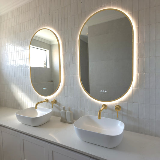 Two Marseille pill-shaped LED mirrors with gold wrap-around frames above twin vessel basins with brushed brass tapware