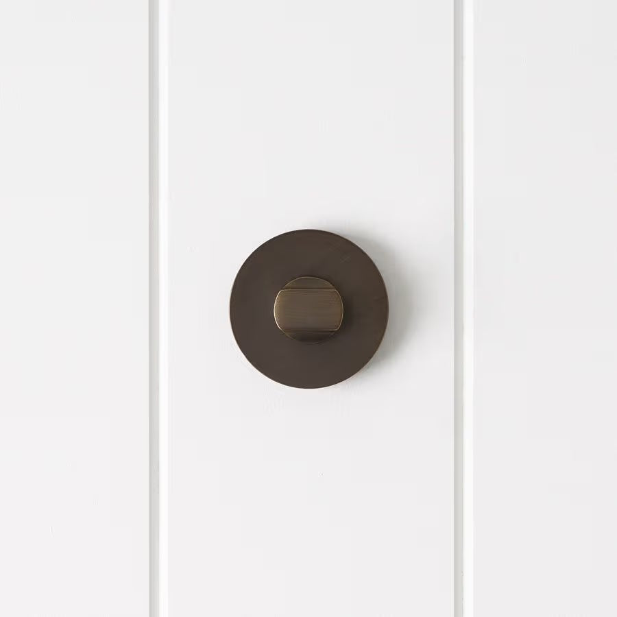 Prague Door Lock in Antique Brass, front view of the thumb-turn on its round rose, white vertical-panelled wall