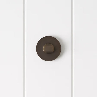 Prague Door Lock in Antique Brass, front view of the thumb-turn on its round rose, white vertical-panelled wall