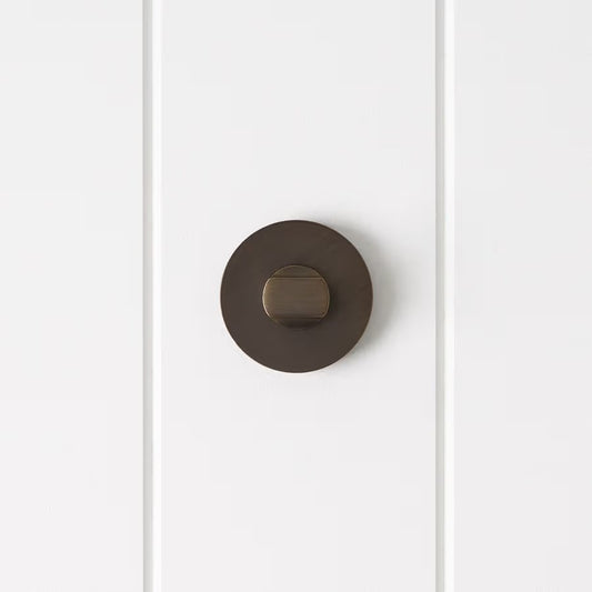 Prague Door Lock in Antique Brass, front view of the thumb-turn on its round rose, white vertical-panelled wall