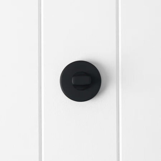 Prague Door Lock in Matte Black, front view of the thumb-turn on its round rose, white vertical-panelled wall