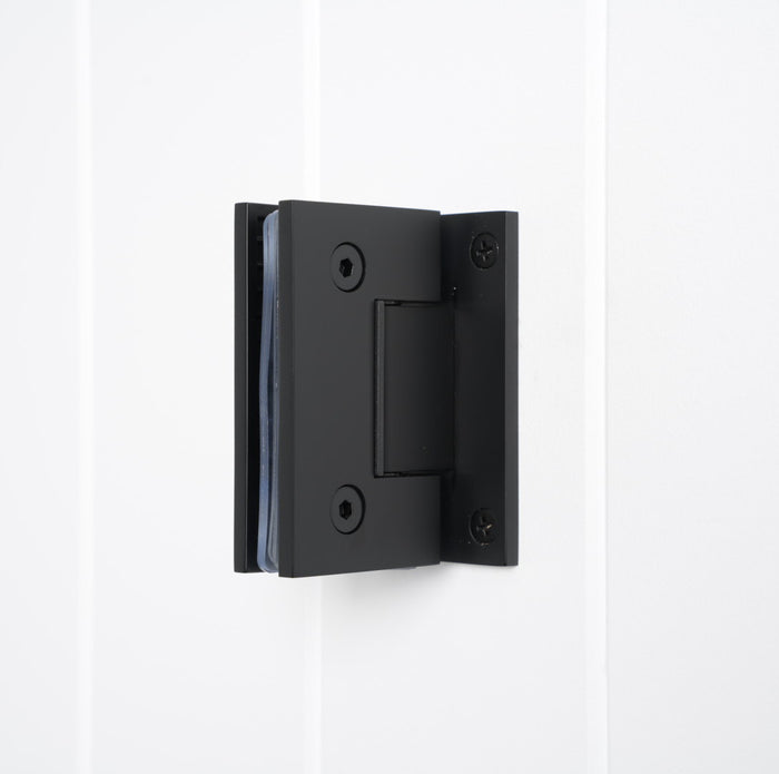 Rennes Shower Hinge Glass to Wall in Matte Black, three-quarter view with clamped glass on a white vertical-panelled wall