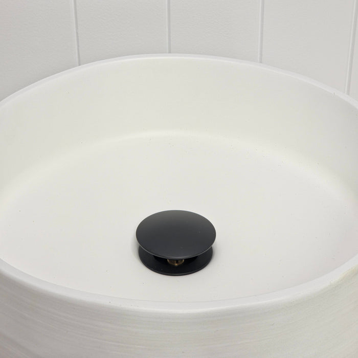 Lisbon Pop Up Basin Waste in Matte Black, plug raised open in a round white basin by a white vertical-panelled wall