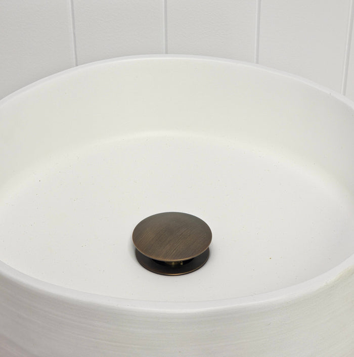 Lisbon Pop Up Basin Waste in Antique Brass, plug raised open in a round white basin by a white vertical-panelled wall